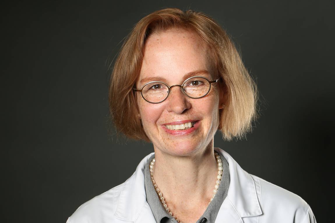 Eine lächelnde Frau mit kurzen hellbraunen Haaren, die eine runde Brille, einen weißen Laborkittel, ein graues Hemd und eine Perlenkette trägt, posiert vor einem schlichten dunklen Hintergrund.