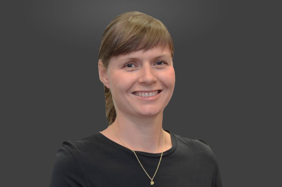 Eine Frau mit zurückgebundenen hellbraunen Haaren, einem schwarzen Oberteil und einer goldenen Halskette lächelt vor einem schlichten dunkelgrauen Hintergrund.